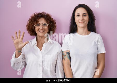 Lateinamerikanische Mutter und Tochter tragen ein legeres weißes T-Shirt auf pinkfarbenem Hintergrund und zeigen mit den Fingern Nummer fünf nach oben, während sie lächeln Stockfoto