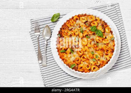 Tortellini gebacken mit Mozzarella-Käse, Tomatensoße und Rinderhack in weißer Backform auf weißem Holztisch mit Löffel und Gabel, horizontale Ansicht Stockfoto