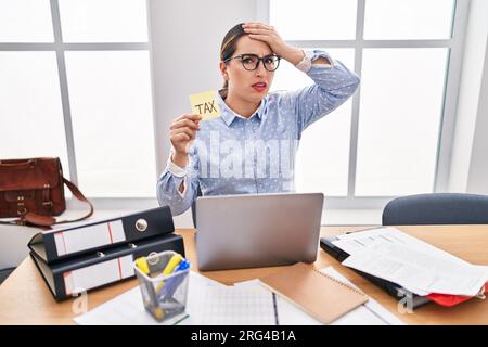 Junge hispanische Geschäftsfrau im Büro, die Steuern kalkuliert, gestresst und frustriert von Hand auf Kopf, überrascht und wütend Stockfoto