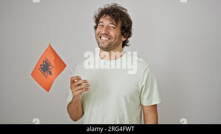 Junger hispanischer Mann, der selbstbewusst lächelt und die albanische Flagge vor einem isolierten weißen Hintergrund hält Stockfoto