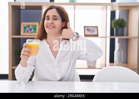 Brünette Frau, die ein Glas Orangensaft trinkt und lächelt und Handygesten mit Hand und Fingern macht, als würde man am Telefon sprechen. Kommunizieren von Konzepten. Stockfoto
