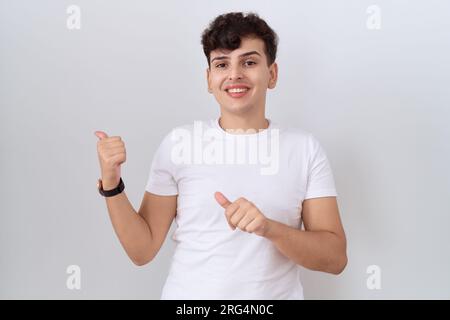 Junger nicht binärer Mann, der ein legeres weißes T-Shirt trägt, das mit der Hand und den Daumen nach oben nach hinten zeigt und selbstbewusst lächelt Stockfoto