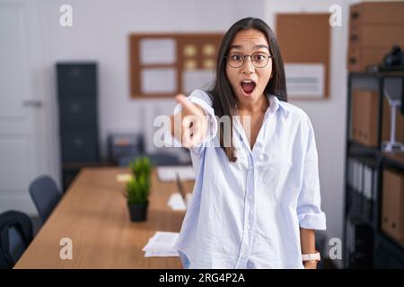 Junge hispanoin im Büro zeigt mit dem Finger überraschend nach vorne, offener Mund, überraschter Ausdruck, etwas auf der Vorderseite Stockfoto
