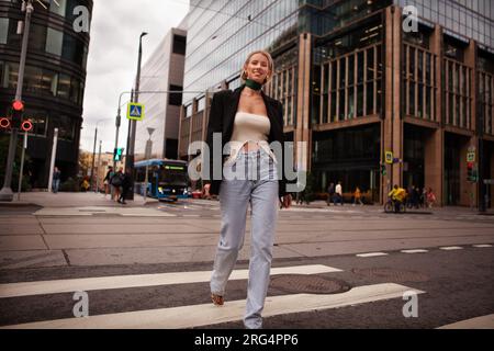 Wunderschöne und stilvolle junge Frau, die die Straße in einer Fußgängerzone in der Stadt überquert, lächelt und modisch in Jeans und Blazer gekleidet ist. Mode Street Style auf BA Stockfoto