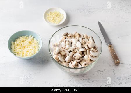 Glasschüssel mit gehackten, frischen Champignon-Pilzen, Schüssel mit geriebenem Käse und gehacktem Knoblauch auf hellblauem Hintergrund mit einem Messer. Phase des Kochens Stockfoto