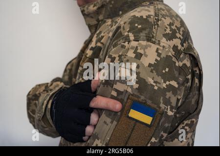 Arm in Handschuh eines ukrainischen Soldaten in Uniform zeigt auf ein Banner der ukrainischen Flagge auf einem Tarnanzug. Pixeliges digitales Soldatenpixel Stockfoto