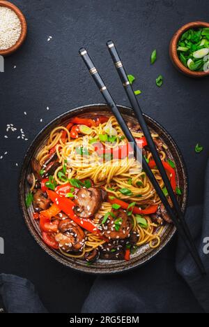 Bratnudeln mit Gemüse umrühren: Rote Paprika, Champignons, grüne Zwiebel und Sesamsamen in Keramikschüssel. Schwarzer Tischhintergrund, Draufsicht Stockfoto