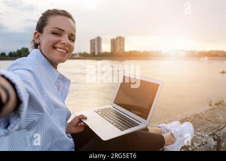 Junge Frau aus der Stadt macht ein Selfie, während sie draußen an einem Laptop vor dem Fluss und der Stadtlandschaft im Hintergrund arbeitet. Stockfoto