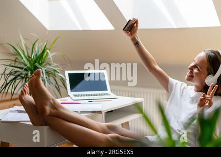Fröhliche junge Frau mit Kopfhörern sitzt zu Hause am Schreibtisch mit erhobenen Füßen und macht ein Selfie mit ihrem Mobiltelefon. Sie filmt sich auf Video. Stockfoto