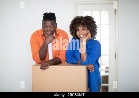 Umzugstag. Schwarze Ehepartner, Die Sich Auf Die Pappschachtel Lehnen Und Die Kamera Anlächeln Stockfoto