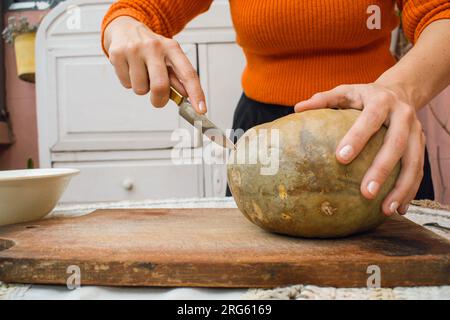 Eine unbekannte Frau zu Hause, die Hallooween einen Kürbis auf einen Tisch schneidet, mit einem Messer, Vorderansicht, Kopierraum. Stockfoto
