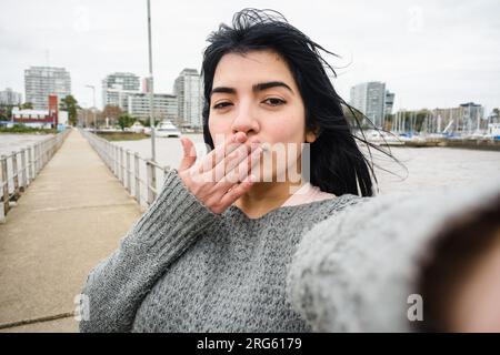 Selfie-Porträt einer jungen, schwarzhaarigen Latina-Frau, die einen grauen Pullover trägt, glücklich lächelt und die Kamera auf dem Pier über dem Fluss im Hafen ansieht Stockfoto