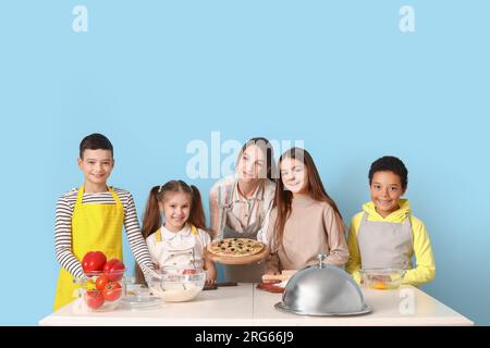 Weiblicher Koch mit zubereiteter Pizza und eine Gruppe kleiner Kinder während des Kochkurses auf blauem Hintergrund Stockfoto