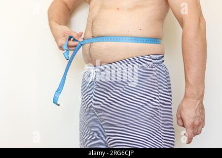 Ein Mann misst seinen fetten Bauch mit Maßband. Konzept der Gewichtsabnahme, Gesundheitsprobleme von übergewichtigen Menschen. Schlechte Ernährung und sitzender Lebensstil. Inkr Stockfoto