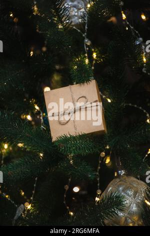 Auf den Zweigen eines grünen Weihnachtsbaums liegt ein Neujahrsgeschenk in handgefertigter Verpackung, gefesselt mit Garn. Festlicher Hintergrund mit leuchtender Girlande und Spielzeug. C Stockfoto