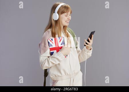 Fröhliches, trendiges Schulmädchen in beigefarbenem Trainingsanzug mit Arbeitsbuch mit englischer Flagge, Kopfhörern und Smartphone gegen Grau. Stockfoto