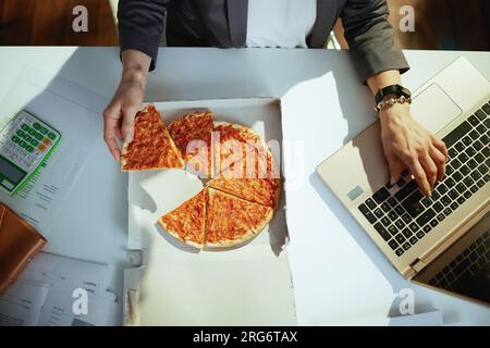 Nachhaltiger Arbeitsplatz. Blick von oben auf die Eigentümerin eines kleinen Unternehmens im grünen Büro mit Pizza und Laptop. Stockfoto