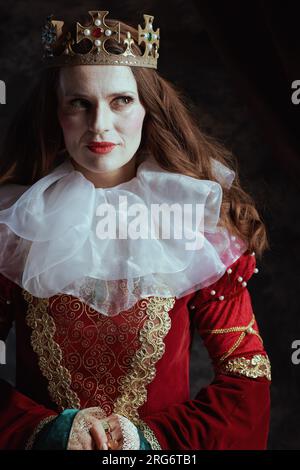 Nachdenkliche mittelalterliche Königin in rotem Kleid mit weißem Kragen und Krone auf dunkelgrauem Hintergrund. Stockfoto