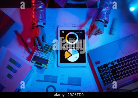 Oberansicht einer modernen Geschäftsfrau mit Taschenrechner, Dokumenten und Laptop, die mit Diagrammen arbeitet und einen Tablet-PC in einem modernen Büro verwendet. Stockfoto