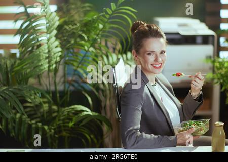 Nachhaltiger Arbeitsplatz. Glückliche, moderne 40-jährige Geschäftsfrau in einem grauen Business-Anzug im modernen grünen Büro, die Salat isst. Stockfoto