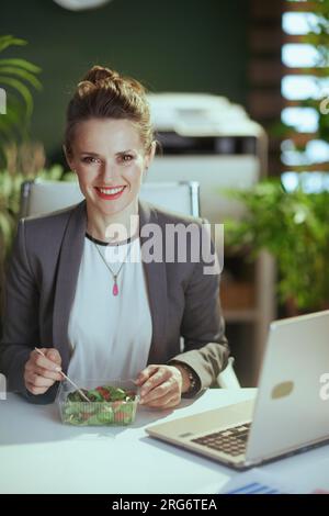 Nachhaltiger Arbeitsplatz. Glückliche, moderne 40-jährige Geschäftsfrau in einem grauen Business-Anzug in einem modernen grünen Büro mit Laptop-Esssalat. Stockfoto