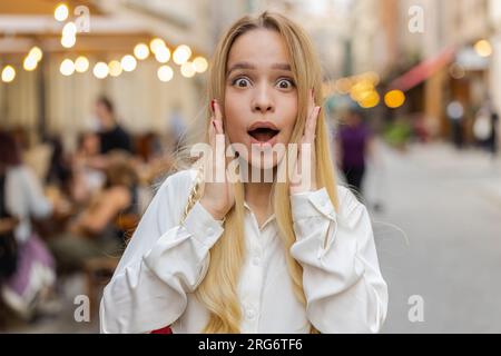 Oh mein Gott, wow. Blonde junge Frau, die überrascht vor der Kamera steht, mit großen offenen Augen, schockiert vom plötzlichen Sieg, Spielsieg, Erreichen von Lotteriezielen, gute Neuigkeiten draußen. Teenager-Mädchen auf der Straße der Stadt Stockfoto