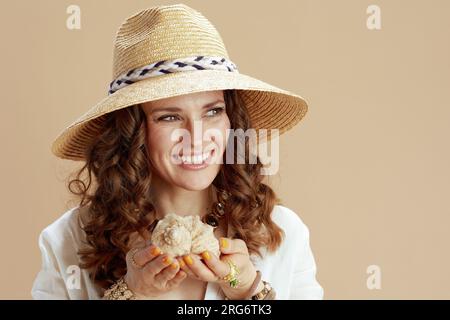 Strandurlaub. Fröhliche, elegante 40 Jahre alte Hausfrau in weißer Bluse und Shorts vor beigefarbenem Hintergrund mit Muschel und Strohhut. Stockfoto