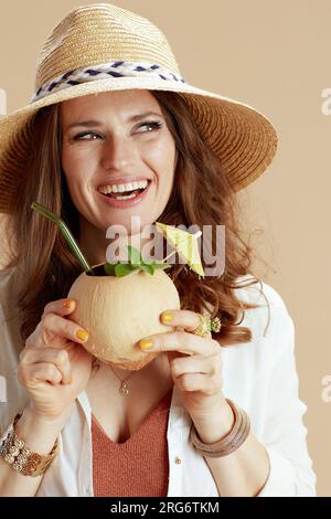 Strandurlaub. Fröhliche, trendige Frau in weißer Bluse und Shorts isoliert auf Beige mit Kokosnuss-Cocktail und Sommerhut. Stockfoto