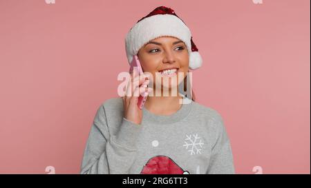 Eine junge Frau im Santa Clause Pullover feiert die Weihnachtsferien und macht Telefongespräche mit Freunden, alles gute. Mädchen, die ihr Smartphone auf einem pinkfarbenen Studiohintergrund unterhalten Stockfoto