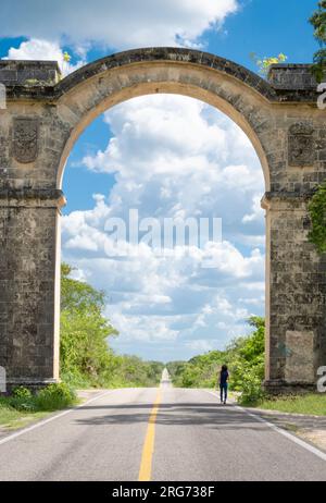 Wunderschöne Landschaft auf einem langen, geraden, leeren Highway, umgeben von Bäumen, der durch einen Steinbogen führt. Eine Frau geht allein auf der Straße Stockfoto