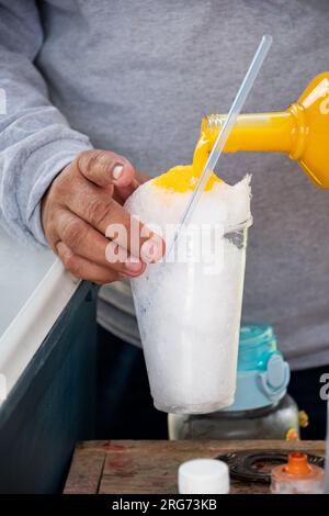 Nahaufnahme eines Lateiners, der Vanille oder Mangosirup in ein Glas mit rasiertem Eis gießt. Snacks, die man auf den Straßen Mexikos findet. Stockfoto