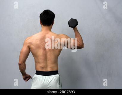 Ein sportlicher Mann hält eine kräftige Kurzhantel hinter seinem Rücken fest und zeigt Stärke und Entschlossenheit. Er steht in einer gut beleuchteten Fitnessatmosphäre, dem pi Stockfoto