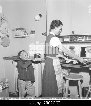 Backen in den 1950er. Eine Mutter mit ihrem Sohn in der Küche. Sie backt süßes Brot und Brötchen. Ein bereits gebackener Teller mit Kochis ist verlockend und der Junge streckt nach einem, wenn seine Mutter wegschaut. Schweden 1958 Kristoffersson Ref BY20-2 Stockfoto