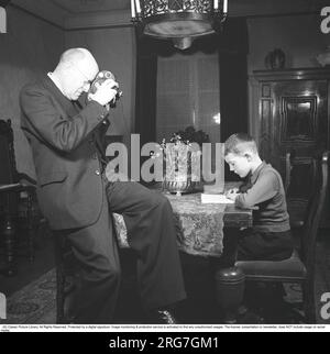 In den 1940er Jahren. Ein Mann filmt mit einer Amateurfilmkamera. Der Film wurde dann entwickelt und auf einem Filmprojektor auf einer klappbaren Leinwand zu Hause gezeigt. Der Film hatte keinen Ton. Die Filmkameras waren nicht batteriebetrieben und man musste sie wie eine Uhr aufziehen, damit sie funktionieren. Er konzentriert sich auf seinen Sohn, der an einem Tisch sitzt und liest. Schweden 1943. Kristoffersson Ref. B103-1 Stockfoto