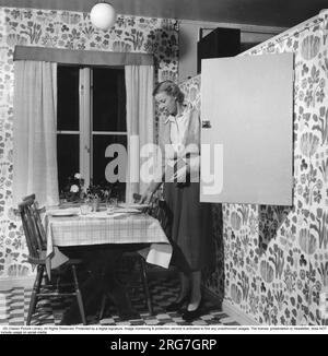 1950er Uhr zu Hause. Eine Frau wird dabei gesehen, wie sie den Tisch für das Abendessen gedeckt hat und die Teller und Gläser, die sie aus einem eingebauten Schrank nimmt, anordnet. Schweden 1950 Stockfoto