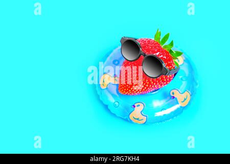 Charmante Figur aus einer frischen, saftigen Erdbeere mit schwarzer Sonnenbrille, schwimmt auf einer aufblasbaren Tube im Pool. Lebhaftes heißes Sommerrefugium Stockfoto