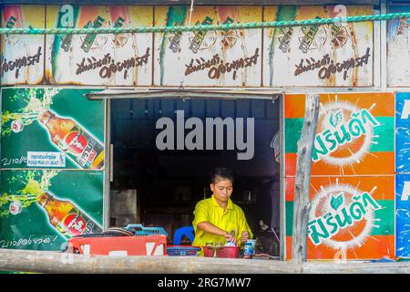 Rayong, Thailand - 4. Dezember 2006: Mann an einem Fenster eines Restaurants reinigt Gerichte und bereitet Essen zu. Die Wand ist mit einem Poster mit alkoholfreien Getränken bedeckt. Stockfoto