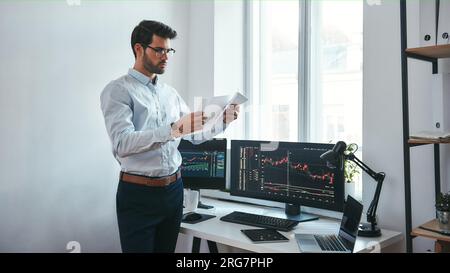 Die Analyse der Daten. Nachdenklich Händler in Brillen Holding einen Finanzbericht und Analyse trading Charts beim Stehen vor Computerbildschirmen in Stockfoto