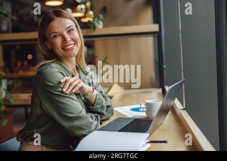 Lächelnde Managerin, die an einem Laptop arbeitet, während sie in gemütlicher Zusammenarbeit am Fenster sitzt Stockfoto