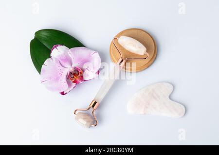 Gua Sha Massagegerät aus Naturstein. Quarzwalze, rosafarbene Orchideenblume, grünes Blatt, weißer Hintergrund, Draufsicht. Gesichtsmassagewerkzeug. Hautpflege, s Stockfoto