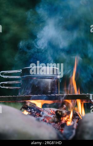Nahaufnahme des kochenden Wassers in einem schwarzen Topf über dem Lagerfeuer auf grünem Hintergrund zur warmen Abendzeit. Lagerfeuer-Kochen in der Sommersaison. Abenteuerkonzept, Stockfoto