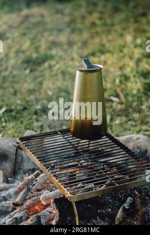 Kaffeezubereitung im Freien am Lagerfeuer im wunderschönen Sonnenaufgang im sommerlichen grünen Park. Campingatmosphäre, Natur- und Outdoor-Lifestyle-Konzept. Lagerfeuer Stockfoto