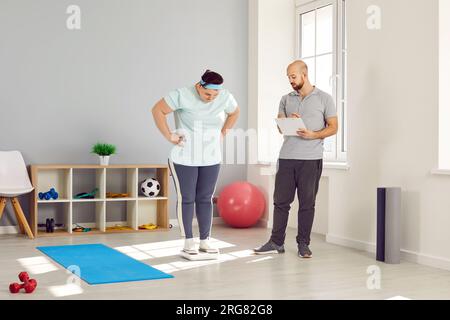 Männlicher Personal Trainer zur Fortschrittskontrolle einer übergewichtigen Frau im Fitnessstudio Stockfoto