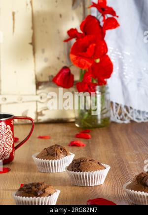 Nahaufnahme des köstlichen Stilllebens von dunklen Schokoladen-Cupcakes, die zu Hause hergestellt werden, umgeben von roten Mohnblüten auf einem rustikalen Holztisch und Details aus Glas Stockfoto