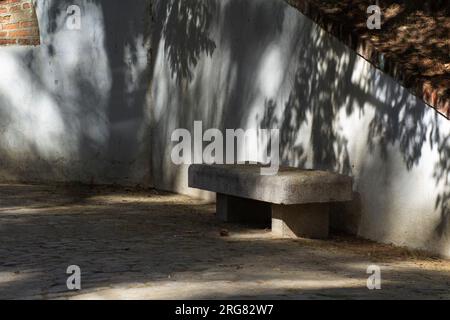 Allgemeine Aufnahme einer einsamen Steinbank in einem Madrider Park im Schatten der Bäume, die auf eine weiße Wand projiziert werden. Horizontale Ansicht. Sonniger Tag. fe Stockfoto