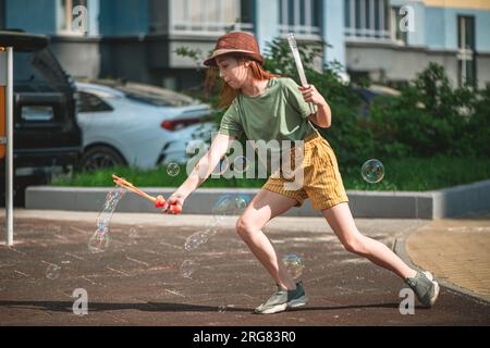 Ein süßes Mädchen im Schulalter bläst im Hof eines mehrstöckigen Gebäudes im Sommer große Seifenblasen. Sommerkonzept, Spiele im Freien Stockfoto