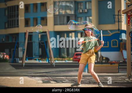 Ein süßes Mädchen im Schulalter bläst im Hof eines mehrstöckigen Gebäudes im Sommer große Seifenblasen. Sommerkonzept, Spiele im Freien Stockfoto