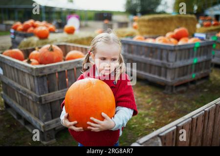 Süßes Vorschulmädchen in rotem Poncho, das Kürbis auf dem Bauernhof auswählt. Fröhliches Kind feiert Halloween Stockfoto