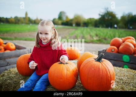 Süßes Vorschulmädchen in rotem Poncho, das Kürbis auf dem Bauernhof auswählt. Fröhliches Kind feiert Halloween Stockfoto