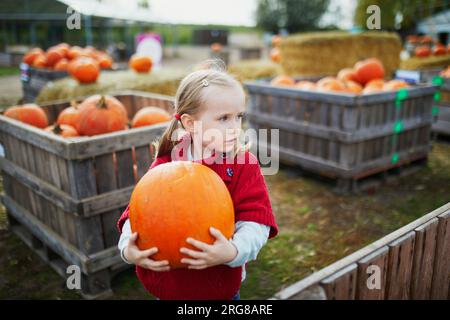 Süßes Vorschulmädchen in rotem Poncho, das Kürbis auf dem Bauernhof auswählt. Fröhliches Kind feiert Halloween Stockfoto